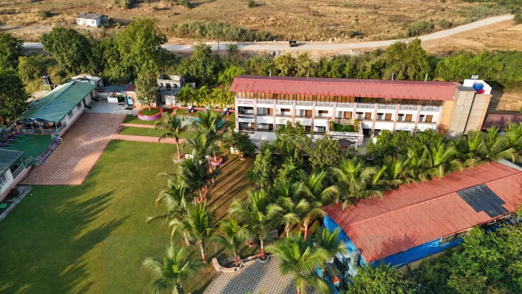 Aerial drone photography of a resort in the Panvel–Karjat region, showcasing the accommodation building, landscaped gardens, palm trees and outdoor recreational areas.